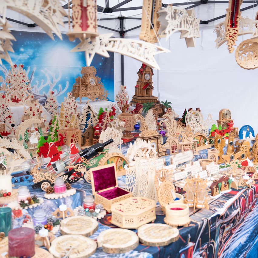 Ein festlicher Marktstand mit kunstvoll geschnitzten Holzdekorationen, Miniaturgebäuden und Weihnachtsbäumen. Verschiedene Gegenstände hängen von der Decke, darunter dekorative Sterne und Uhren.