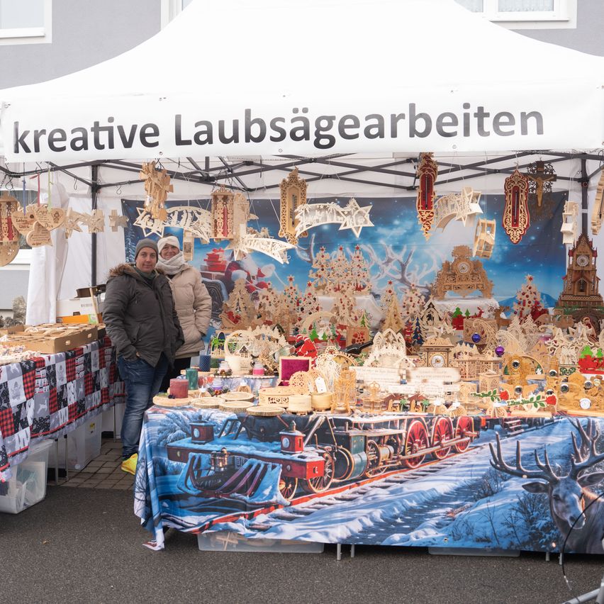 Ein Mann und eine Frau stehen vor einem Stand auf einem Weihnachtsmarkt. Der Stand ist von einem weißen Zelt mit einem Banner bedeckt, auf dem 'kreative Laubsägearbeiten' steht. Der Stand zeigt eine Vielzahl von Holzschmuck und einem Zugmodell.