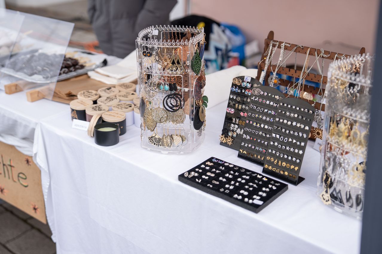 Ein Tisch mit weißem Tuch zeigt verschiedene Schmuckstücke, darunter Ohrringe, Halsketten und Ringe, auf Ständern und in Gläsern angeordnet.
