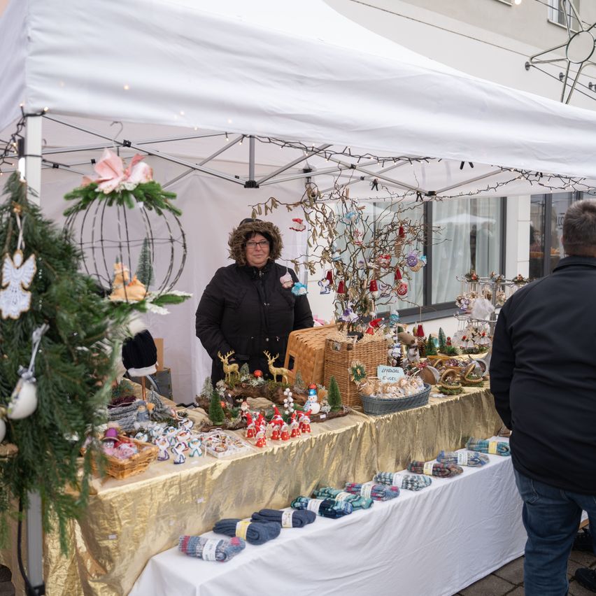 Eine Frau steht unter einem weißen Zelt und verkauft Weihnachtsartikel. Verschiedene Artikel sind auf einem goldenen Tisch ausgestellt, darunter Socken, Ornamente und Körbe. Ein Mann steht in der Nähe.