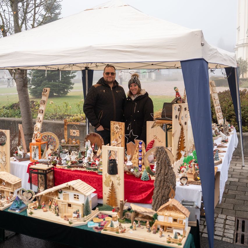 Zwei Personen stehen hinter einem Tisch mit Holzornamenten. Sie stehen vor einem weißen Zelt und einer Kirche.