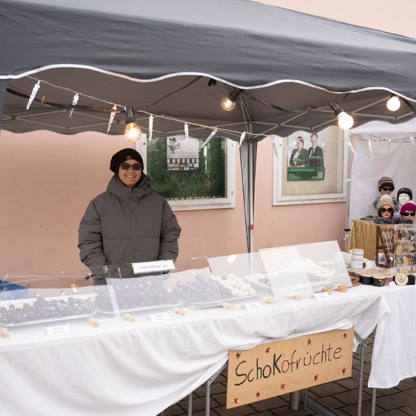 Eine Frau steht hinter einem Tisch unter einem Zelt und verkauft Schokoladen. Ein Schild auf dem Tisch steht 'SchoKofrüchte'. Das Zelt ist mit Lichtern geschmückt. Hinter dem Tisch befinden sich zwei weitere Tische mit Gegenständen darauf.