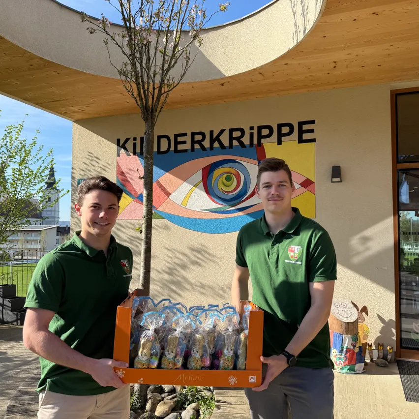 Zwei Männer in grünen Polo-Shirts halten eine Box mit Ostereiern vor einem Gebäude mit dem Schild 'Kil DERKriPPE'.