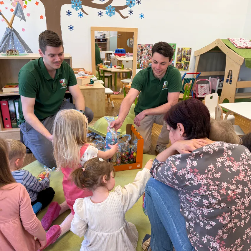 Zwei Männer in grünen Poloshirts knien auf dem Boden in einem Spielzimmer und verteilen Süßigkeiten an eine Gruppe von Kindern, die in einem Kreis sitzen.