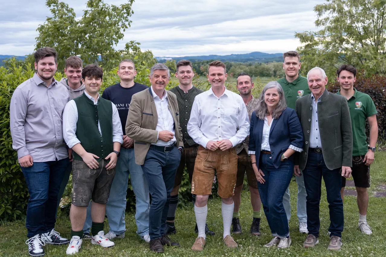 Eine Gruppe von Personen in traditioneller Kleidung steht auf einem Rasen. Die Männer tragen grüne Westen und braune Shorts, während die Frau einen blauen Blazer trägt. Sie lächeln und posieren für ein Foto.