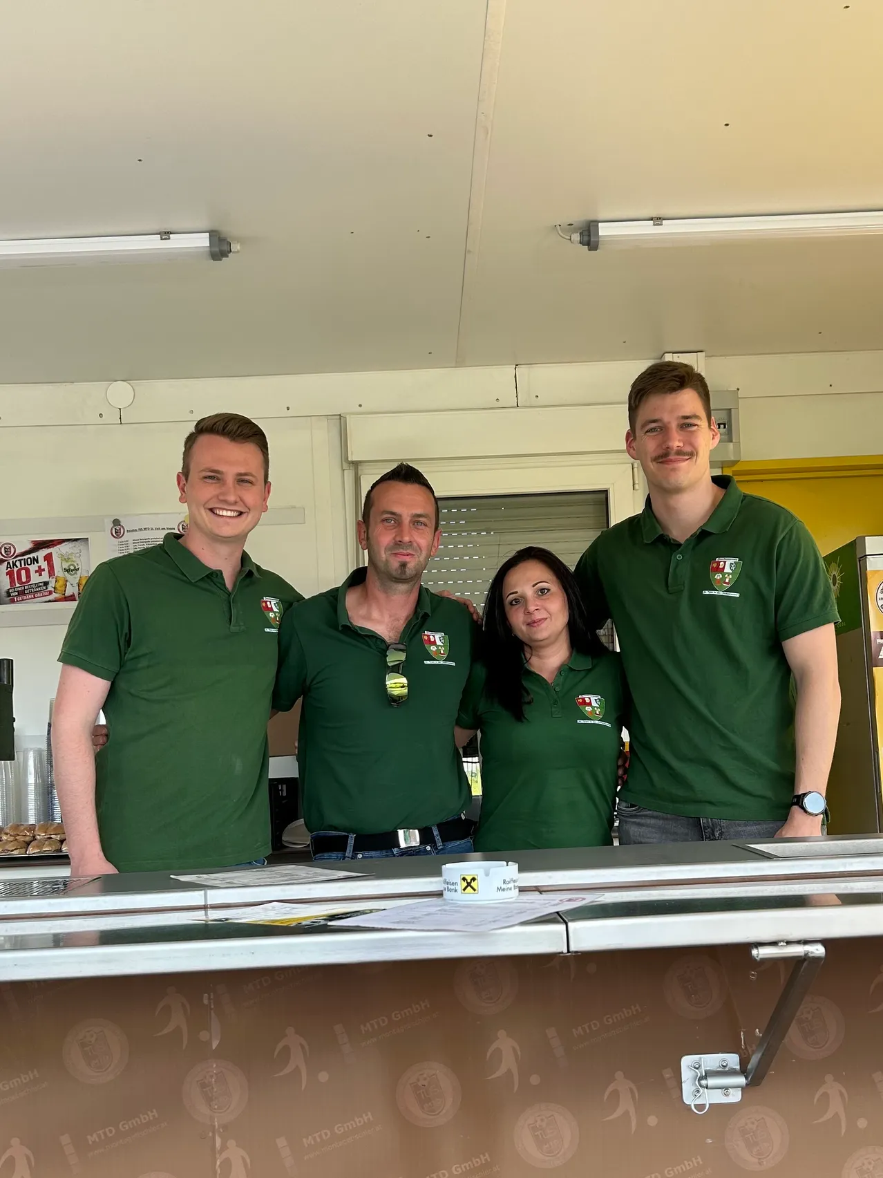 Vier Menschen stehen lächelnd für ein Foto vor einem Tisch. Sie tragen passende grüne Poloshirts mit Logo. Dahinter befinden sich Regale mit Essen, Becher und ein Plakat. Oben ist eine helle Lampe und ein offenes Fenster.