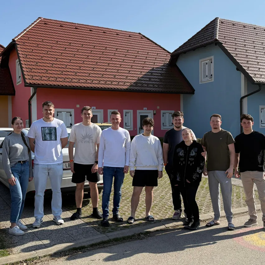 Eine Gruppe von Menschen steht vor zwei Häusern, eines rot und eines blau, mit einem Auto davor.
