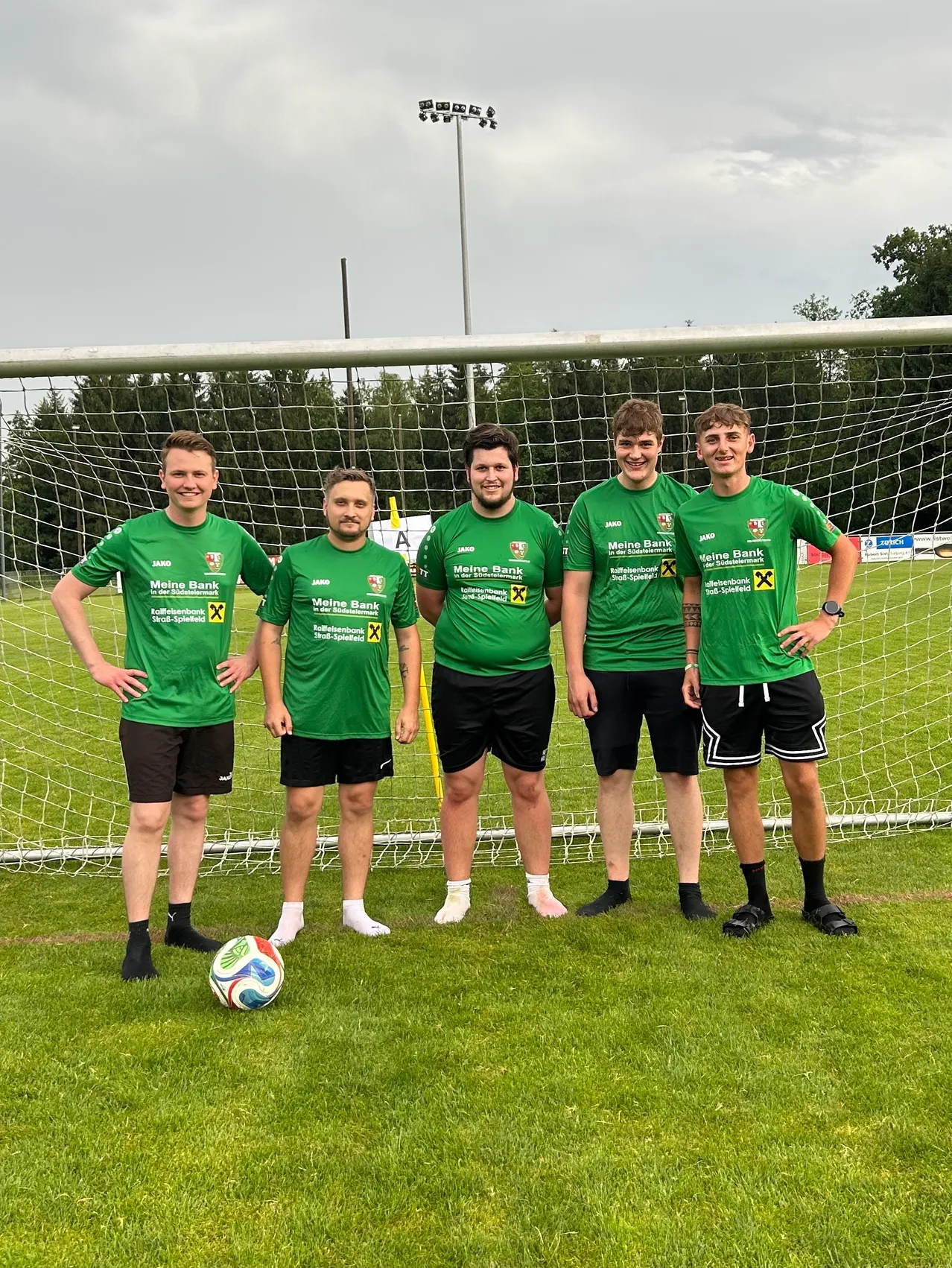 Fünf Männer in grünen Trikots mit einem Fußball vor einem Netz. Sie lächeln und posieren für ein Foto.