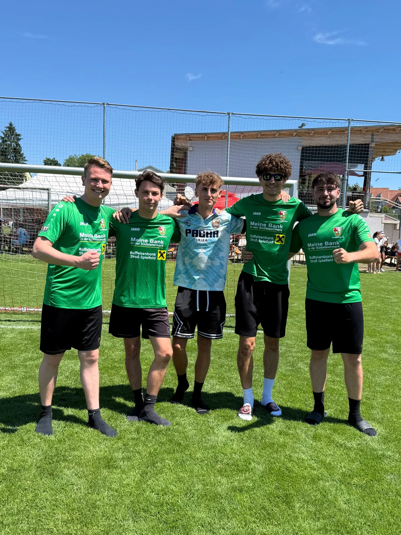 Fünf junge Männer in grünen Trikots mit 'Meine Bank' posieren für ein Bild auf einem Fußballfeld mit einem Tor und einem Gebäude im Hintergrund.