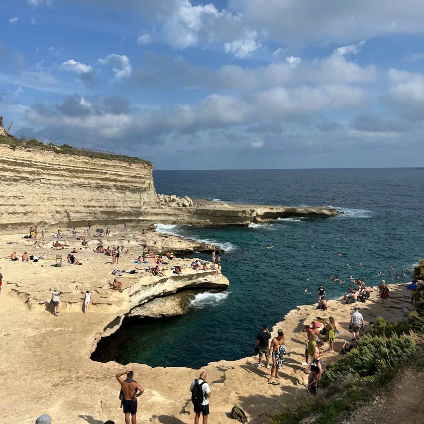 Eine Gruppe von Menschen genießt einen sonnigen Tag an einem Strand am Fels. Einige sonnen sich, während andere im klaren blauen Wasser schwimmen.