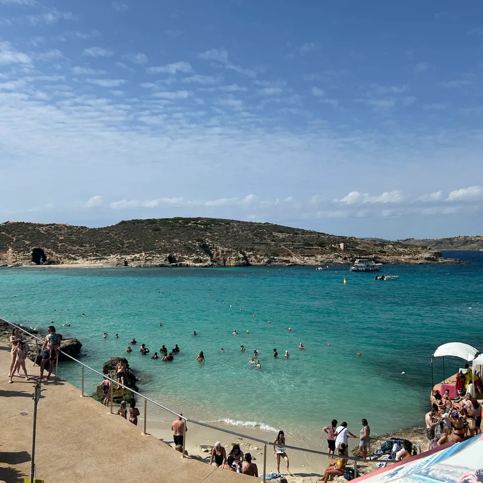 Ein Strand mit vielen Menschen, die im blauen Wasser schwimmen. In der Ferne sind Boote zu sehen, und eine kleine Insel ist sichtbar.
