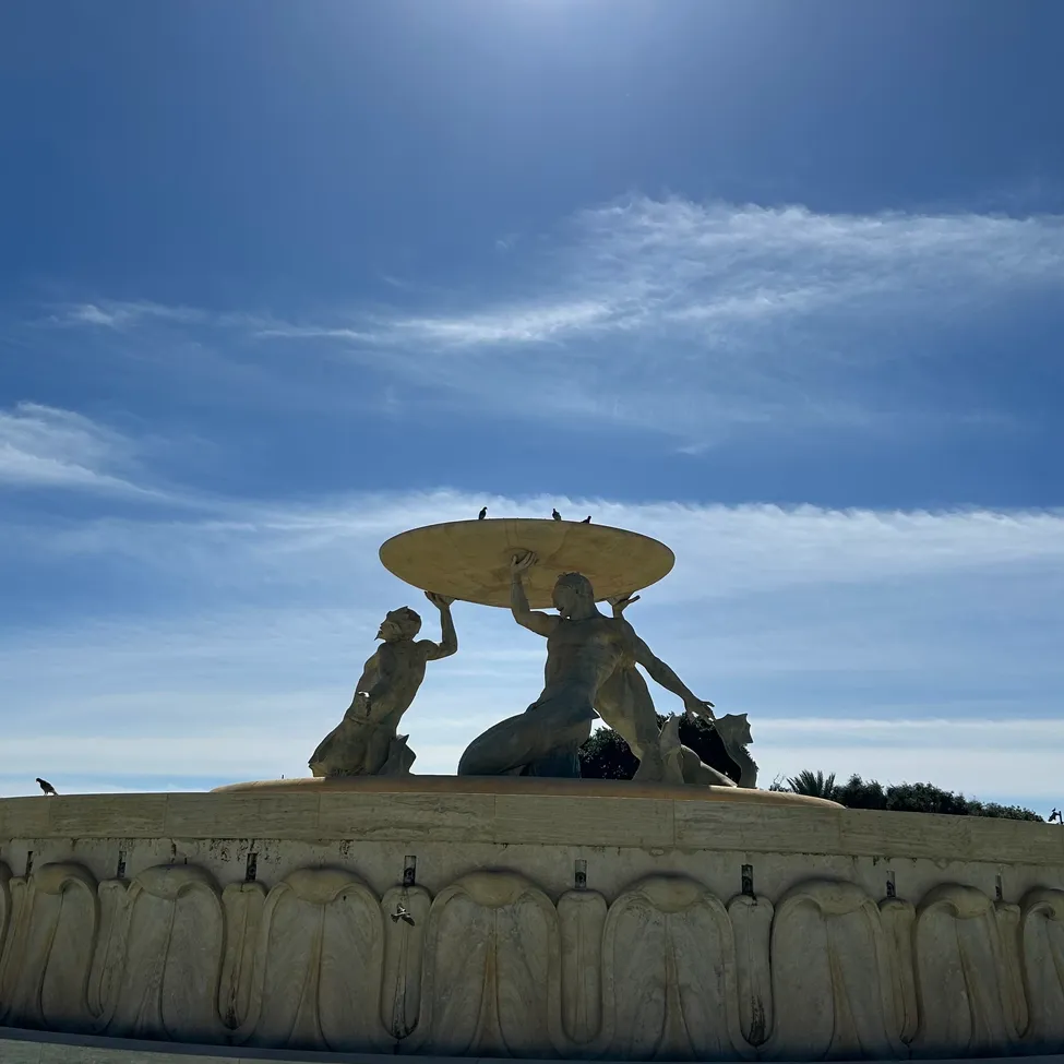 Eine Skulptur von zwei Personen, die eine große Scheibe unter einem hellen Sonnenschein gegen einen blauen Himmel mit Wolken halten.