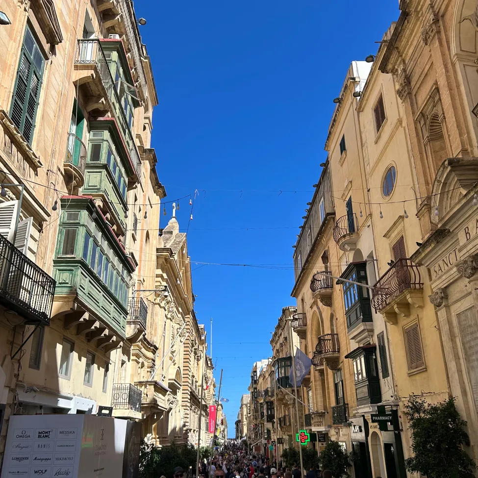 Eine belebte Straße in einer Stadt mit vielen Menschen, die auf dem Bürgersteig gehen. Gebäude mit Balkonen und Fenstern säumen die Straße, und der Himmel ist klar und blau.