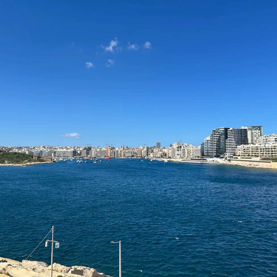 Ein Panoramablick auf eine Küstenstadt mit einem großen Gewässer, mehreren Gebäuden und einem klaren blauen Himmel.
