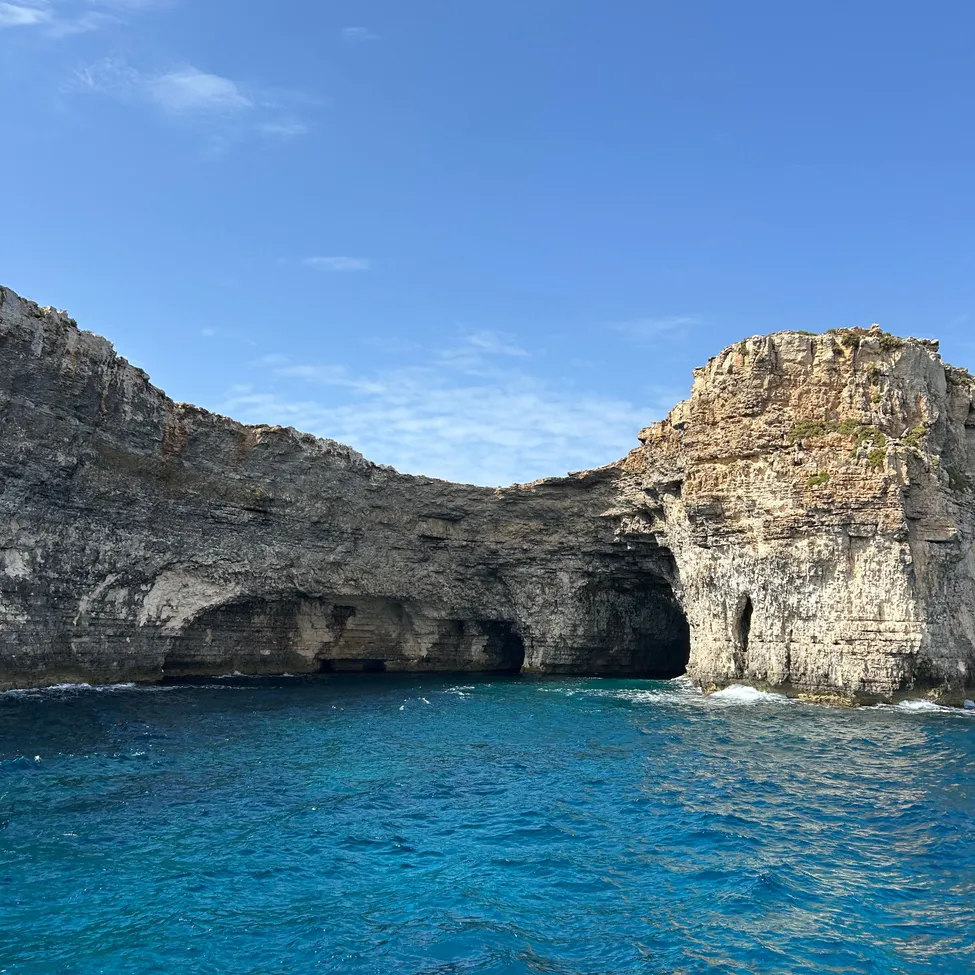 Eine große Felsformation mit einer Höhlenöffnung in der Mitte, umgeben von blauem Wasser unter einem klaren blauen Himmel.