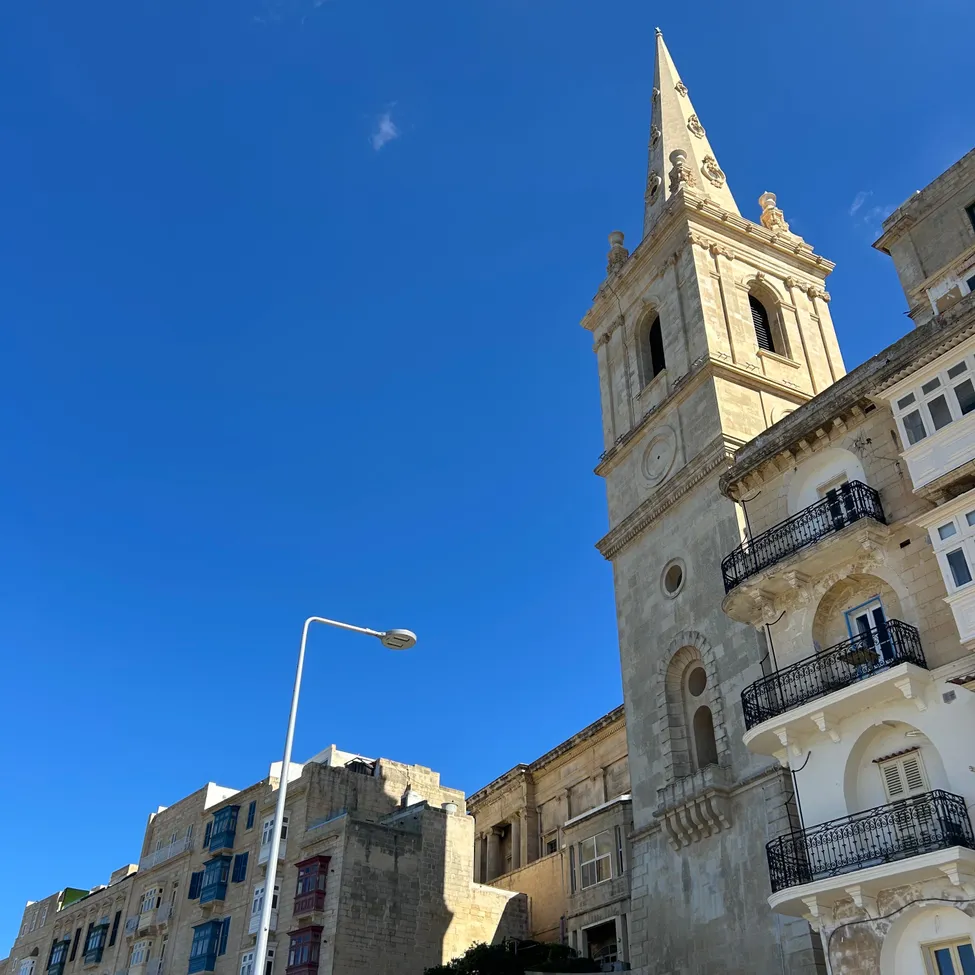 Ein hoher, verzierter Turm steht vor einem großen Gebäude mit Balkonen. Der Turm ist aus Stein und hat eine spitze Spitze. Der Himmel ist blau mit ein paar Wolken.