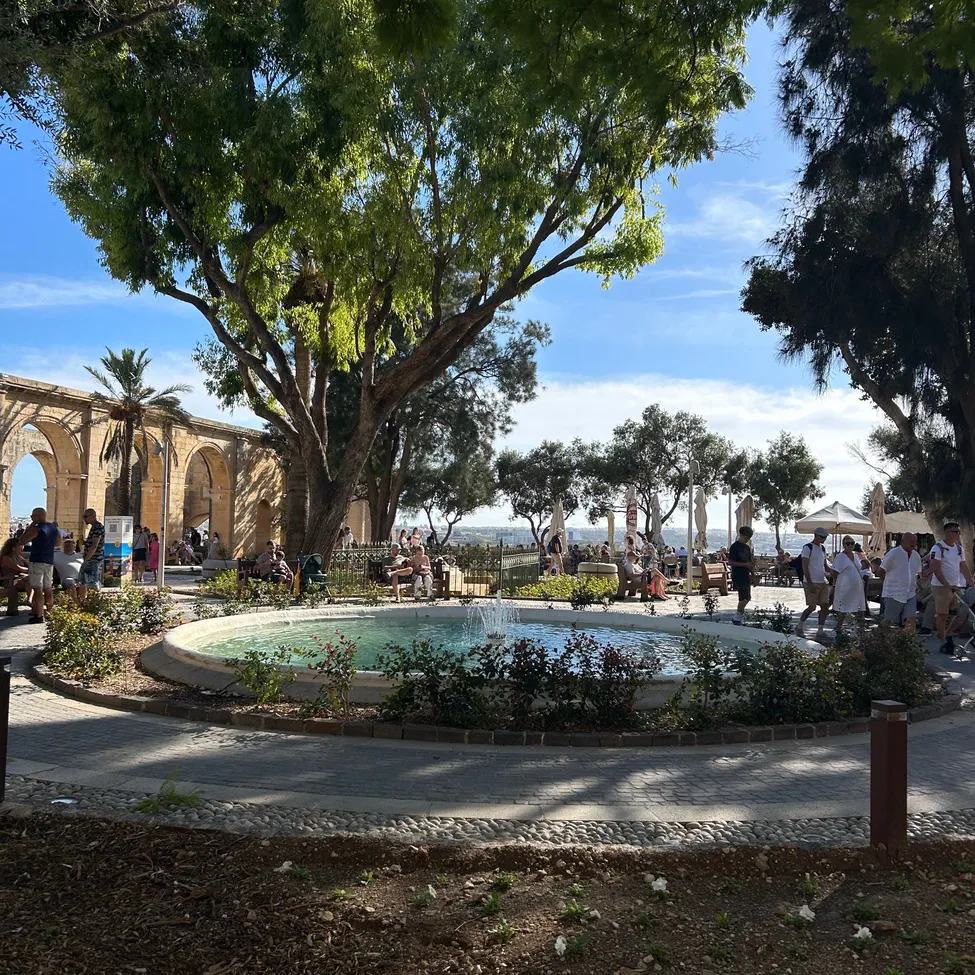 Ein Park mit einem Brunnen in der Mitte, umgeben von Bäumen und Menschen, die sitzen und gehen.