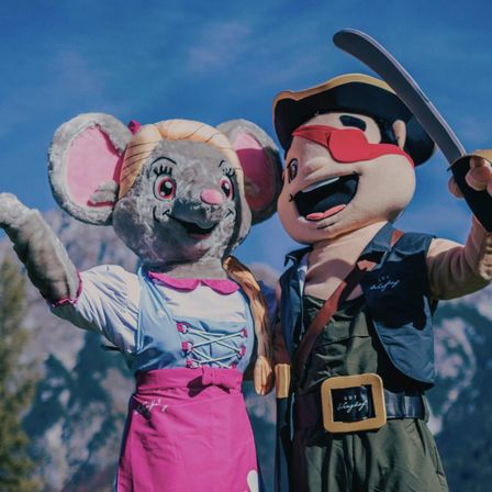 Zwei Maskottchen, eine Maus und ein Pirat, stehen nah beieinander vor einer Bergkulisse.