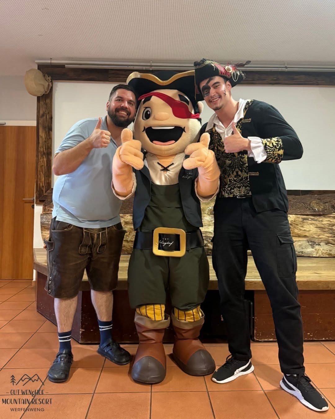 Zwei Männer und ein Piratenmaskottchen posieren für ein Foto, alle lächeln und geben Daumen hoch. Das Maskottchen trägt einen Piratenhut und einen Gürtel, während die Männer in lässiger Kleidung sind.