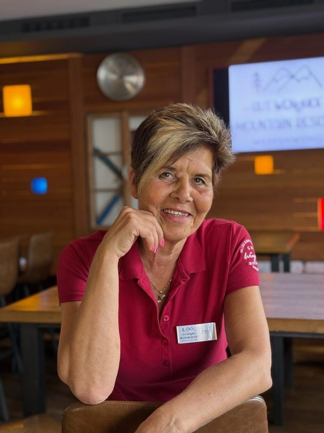 A woman with short blonde hair and a red polo shirt sits at a table in a restaurant. She is smiling and has her hand on her chin.