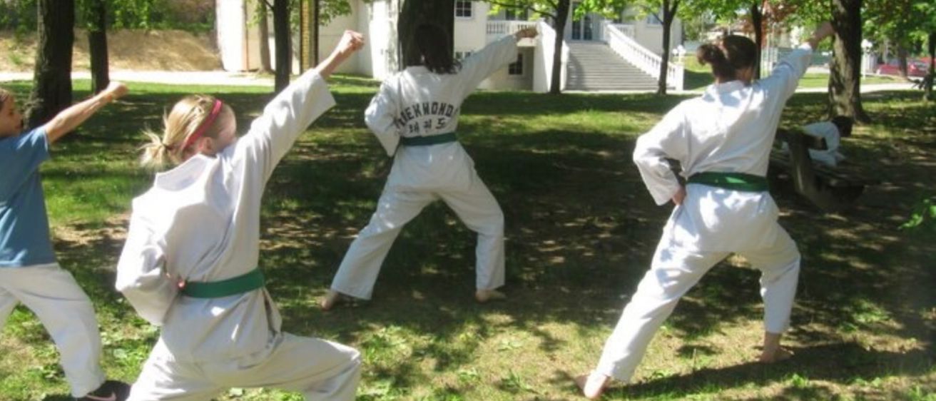 Drei Personen in weißen Kampfsportuniformen üben Karate in einem Park mit einem großen Baum und einem Gebäude mit Treppen im Hintergrund.