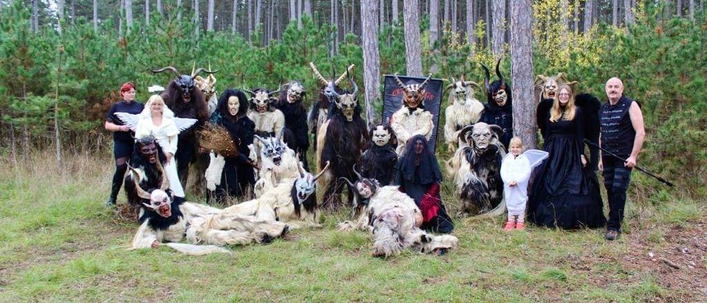 Eine Gruppe von Menschen in Kostümen, einige mit Hörnern, Masken und Flügeln, posiert für ein Foto in einem Wald. Hohe Bäume umgeben das Gebiet.