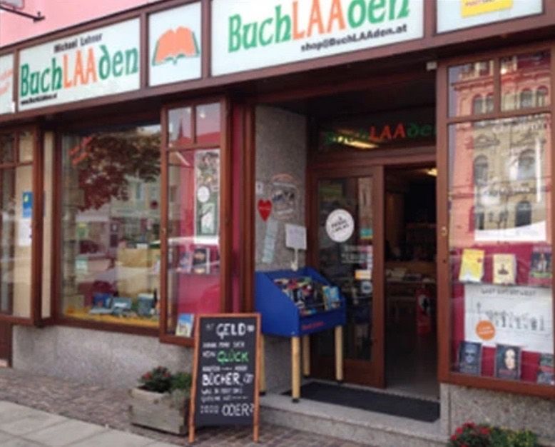 Ein Buchladen namens BuchLAAden mit einem Schild über dem Eingang. Ein Schwarzes Brett am Bürgersteig bewirbt einen schnellen Geldwechselservice.