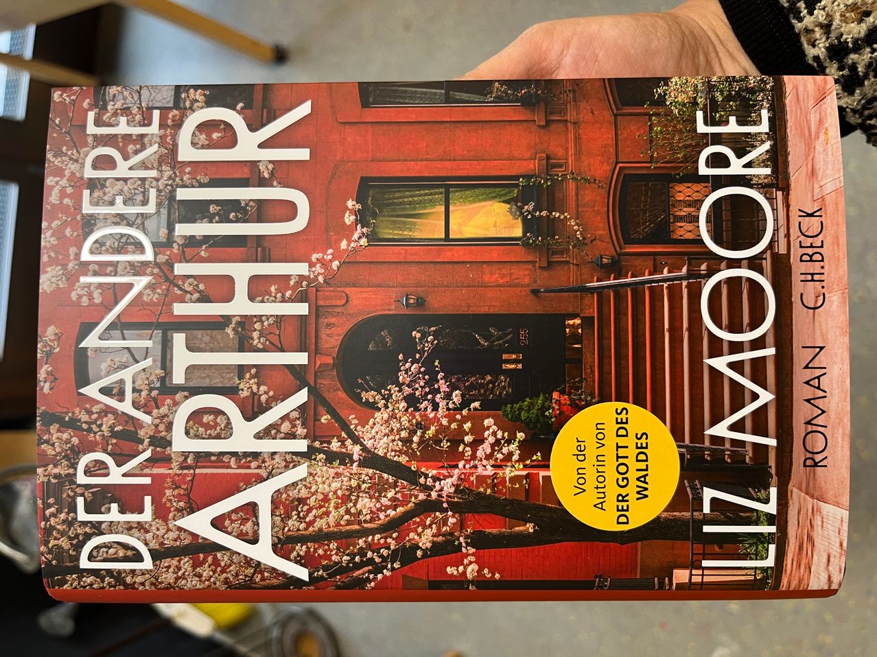 Buchcover für 'Arthur' von DeRandeere, zeigt ein rotes Gebäude, einen Baum mit rosa Blüten und einen gelben Kreis mit deutschem Text.