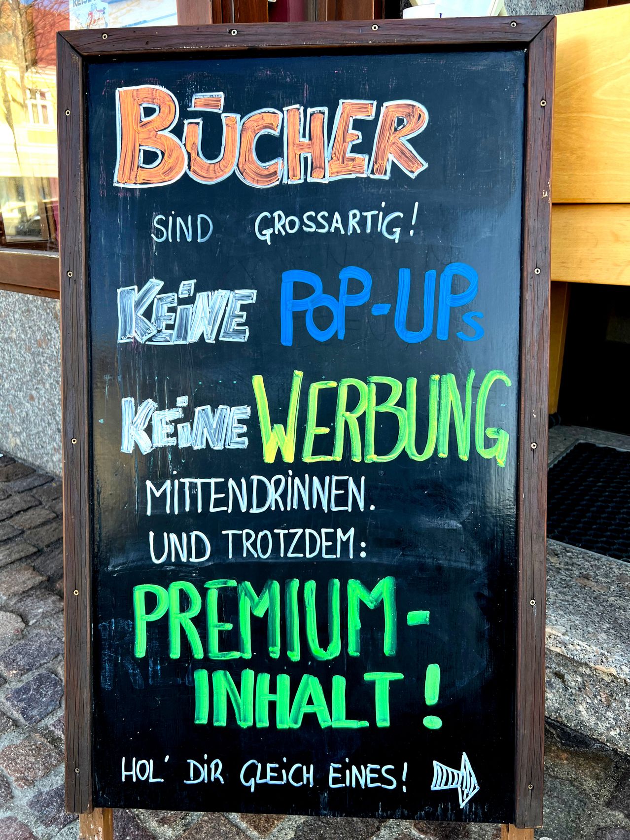 Ein Kreidetafel-Schild vor einem Gebäude bewirbt Bucher. Es steht 'Bucher sind großartig!', 'Keine Pop-Ups', 'Keine Werbung', 'Keine Zwischengetränke' und 'Trotzdem: Premium-Inhalt!'.