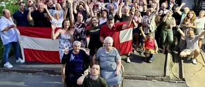 Eine Gruppe von Menschen steht zusammen und hält eine Flagge. Viele heben ihre Hände, und einige tragen Brillen und Hüte. Eine Person hält eine Flagge in der Mitte, während eine andere eine Flasche hält. Eine rote Flagge ist links, und einige Leute sitzen auf Stühlen davor.