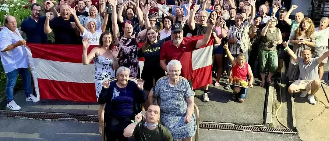 Eine Gruppe von Menschen steht zusammen und hält eine Flagge. Viele heben ihre Hände, und einige tragen Brillen und Hüte. Eine Person hält eine Flagge in der Mitte, während eine andere eine Flasche hält. Eine rote Flagge ist links, und einige Leute sitzen auf Stühlen davor.