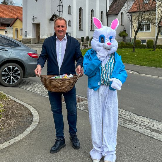 Ein Mann in einem marineblauen Anzug und eine Person in einem Hasenkostüm halten einen Korb voller Gegenstände. Sie stehen auf der Straße vor einer Kirche.