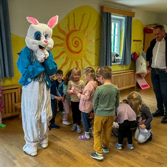 Ein Erwachsener, der als Osterhase verkleidet ist, gibt Kindern, die sich in einer Reihe aufstellen, um sie zu erhalten, Süßigkeiten. Dahinter befindet sich eine Wand mit einem Sonnendesign. Daneben steht ein Mann neben einer Tasche, die an einem Haken hängt.