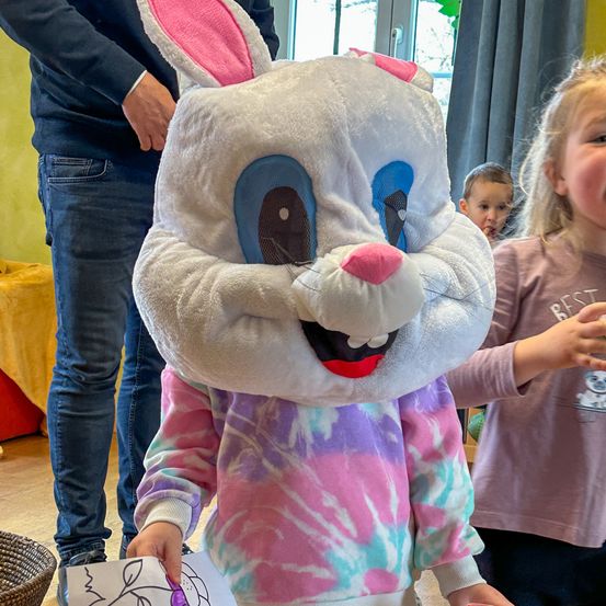 Ein junges Kind in einem Plüschhasenkostüm hält eine Papierblume und steht neben zwei anderen Kindern.