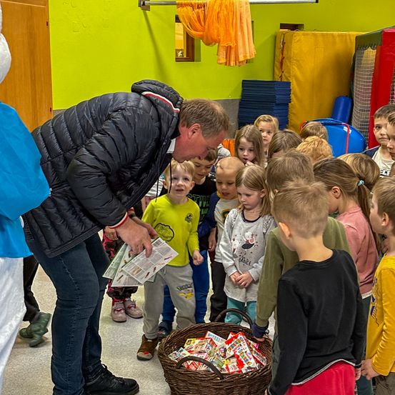 Ein Mann in Schwarz verteilt Süßigkeiten an eine Gruppe junger Kinder in einem Klassenzimmer.