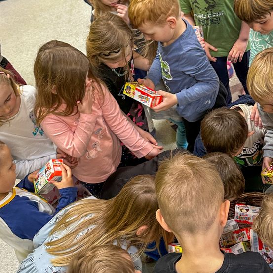 Eine Gruppe von Kindern ist versammelt, einige halten Boxen, wahrscheinlich in einem Klassenzimmer.