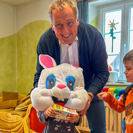 Ein Mann hält einen großen Plüschhasen, während ein junger Junge eine Schachtel mit Süßigkeiten in der Hand hat. Der Junge trägt ein T-Shirt mit einem Hasen-Design. Ein zweiter Junge, der ein orangefarbenes Shirt trägt, steht neben ihnen.