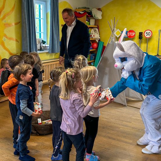 Ein Maskottchen in einem Hasenkostüm gibt Kindern Geschenke in einem Raum mit gelber Wand, STOP-Schild und einem Korb auf dem Boden.