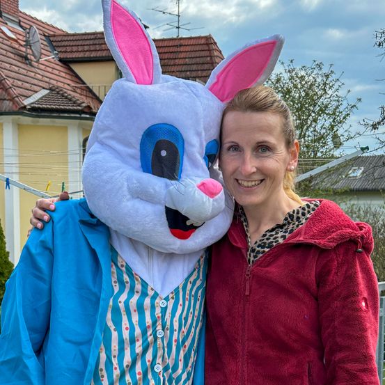 Eine Frau in einer roten Jacke und ein Mann in einer blauen Jacke und Hasenkostüm posieren für ein Foto vor einem Haus mit rotem Dach.