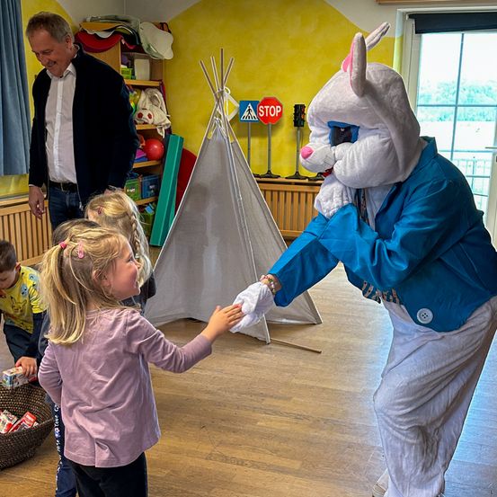 Ein Mann im Anzug steht hinter einem kleinen Mädchen, das mit einer Osterhasen-Maskottchen die Hand schüttelt.