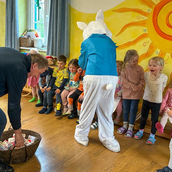 Ein Mann, verkleidet als Osterhase, verteilt Ostereier an eine Gruppe von Kindern, die im Kreis sitzen. An der Wand hinter ihnen ist eine große Sonne gemalt.