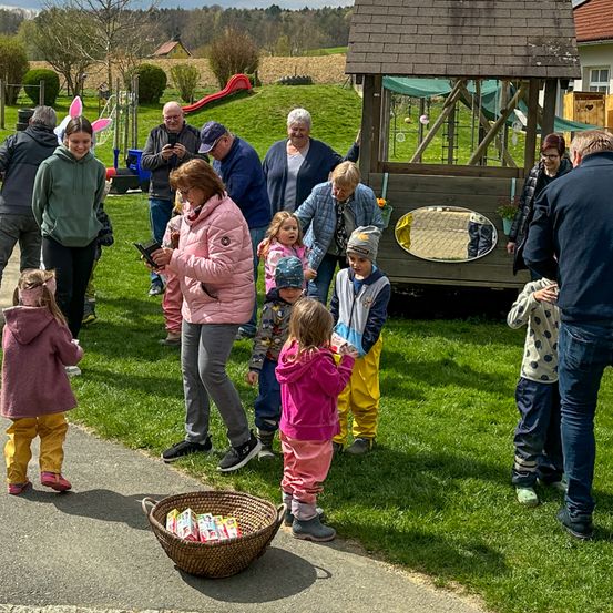 Eine Gruppe von Menschen versammelt sich in einem Außenbereich, möglicherweise einem Park, mit einem Korb voller Schokoladeneiern auf dem Boden. Es gibt Kinder und Erwachsene in der Gruppe, und einige von ihnen tragen Mützen und Brillen. Hinter ihnen steht ein hölzerne Spielhaus mit einem Spiegel und einer Topfpflanze.