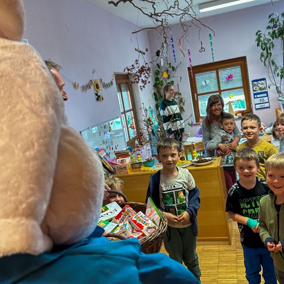 Eine Gruppe von Kindern steht in einem Klassenzimmer mit einer Lehrerin, umgeben von einem dekorierten Raum. Einige halten Spielzeug, andere lächeln, während eine Person in einem blauen Hemd einen großen Plüschtier hält. Der Raum ist mit Dekorationen, Pflanzen und einem Holztisch mit Gegenständen darauf gefüllt.