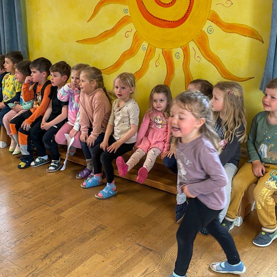 Eine Gruppe von Kindern sitzt in einem Klassenzimmer, mit einem großen Sonnenwandbild an der Wand.