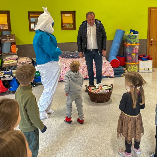 Ein Mann und eine Maskottchen stehen neben einem Korb mit Süßigkeiten. Eine Gruppe von Kindern steht um sie herum, und eines der Kinder schaut in den Korb.