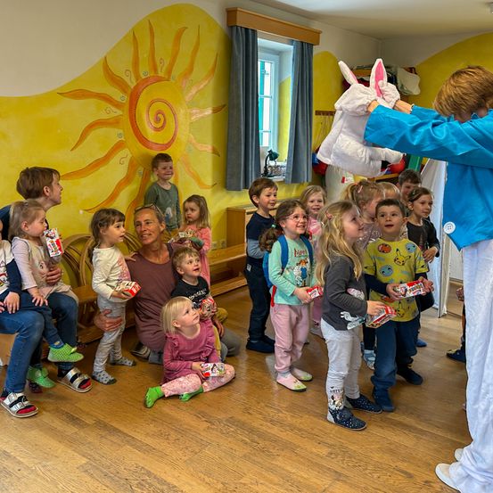 Eine Gruppe von Kindern und Erwachsenen in einem Raum, einige sitzen auf dem Boden, andere stehen, und eine Frau in einer Hasenkostüm verteilt Spielzeug. Der Raum hat eine Sonne an der Wand und eine Bank.