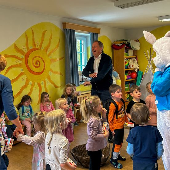 Eine Gruppe junger Kinder steht in einem Kreis in einem Spielzimmer mit einem Mann als Osterhase verkleidet und einem Mann in einem schwarzen Mantel, der sie anlächelt.