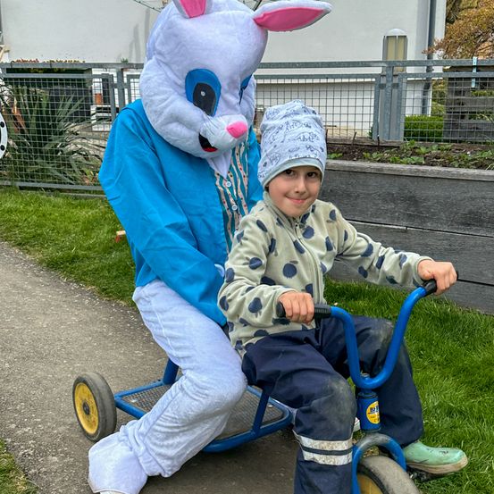 Ein Junge fährt auf einem Dreirad, während eine Person als Osterhase die Lenkstange hält.