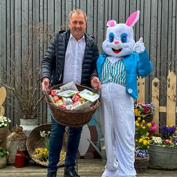 Ein Mann in einer schwarzen Jacke hält einen Korb mit Ostergeschenken und steht neben einer Person, die als Hase verkleidet ist. Sie lächeln beide, und im Hintergrund befinden sich mehrere Blumentöpfe und Pflanzen.