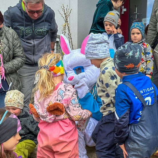 Eine Gruppe von Kindern in Winterkleidung versammelt sich um ein großes weißes Hasenkostüm. Einige der Kinder tragen Mützen, während andere Stirnbänder haben. Ein Mann steht hinter dem Hasenkostüm, und eine Frau hält eine Blume. Die Szene spielt sich vor einem Gebäude mit einer Glastür ab.
